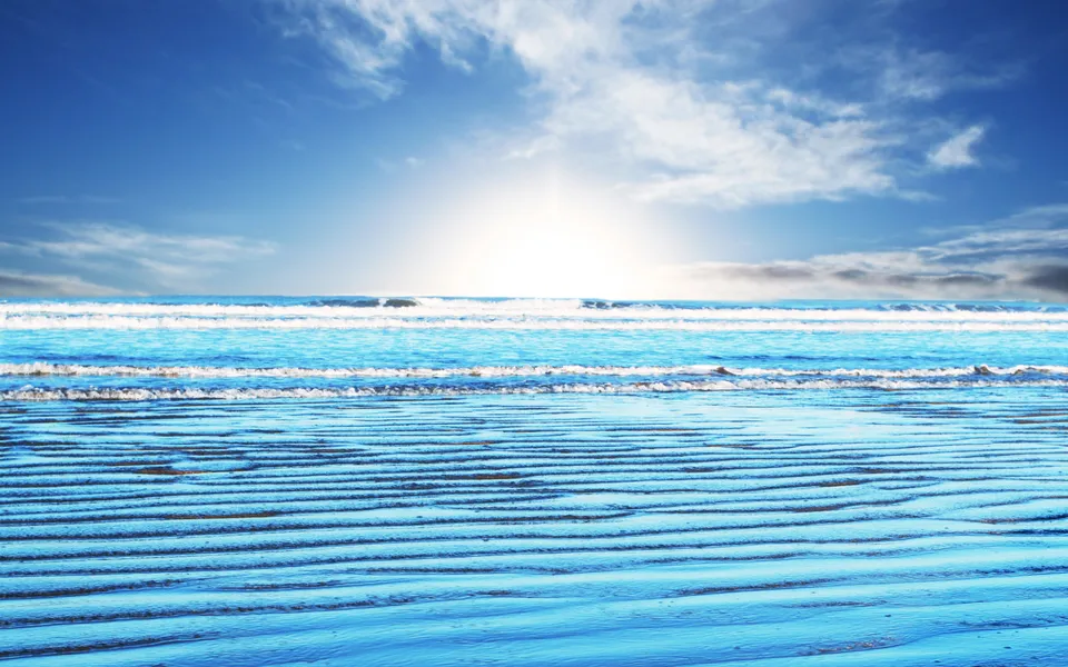 Close-up of rippled wet sand on a beach with gentle waves and a bright sky in the background.