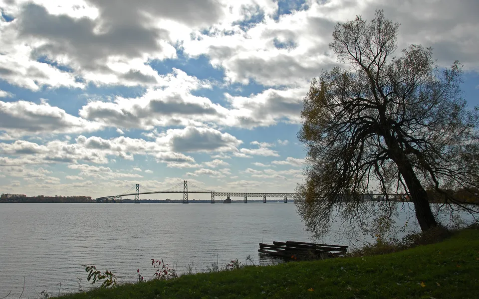 A long suspension bridge crosses a wide river under a sky filled with dramatic clouds.