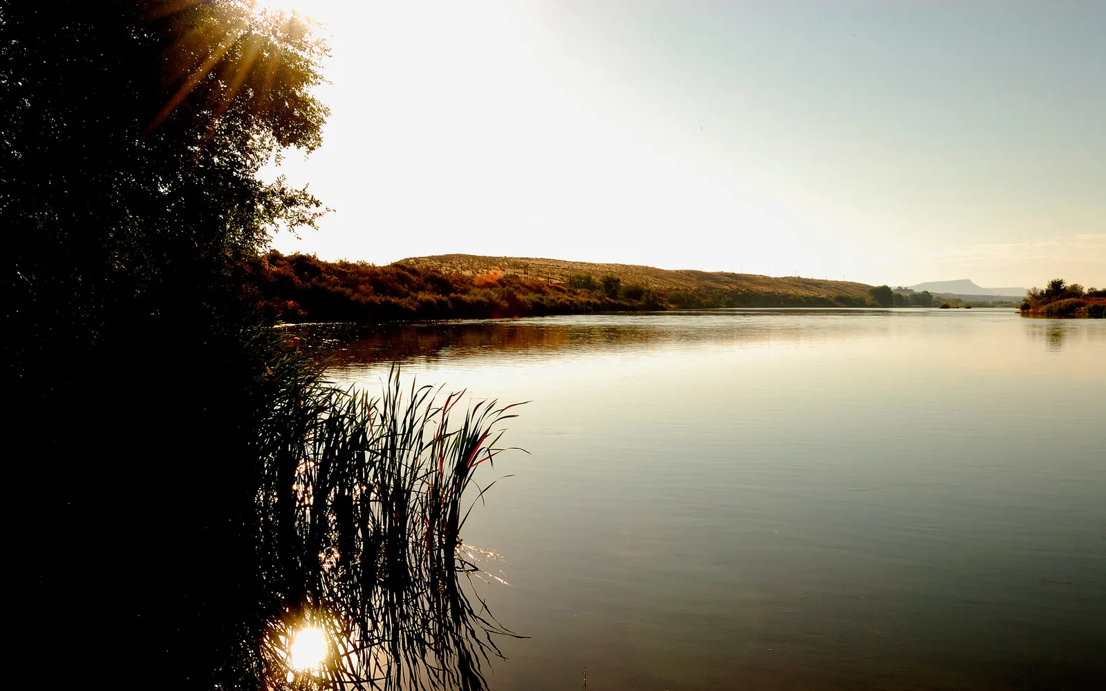 A wide river reflects the bright sun and surrounding hills, with reeds in the foreground.