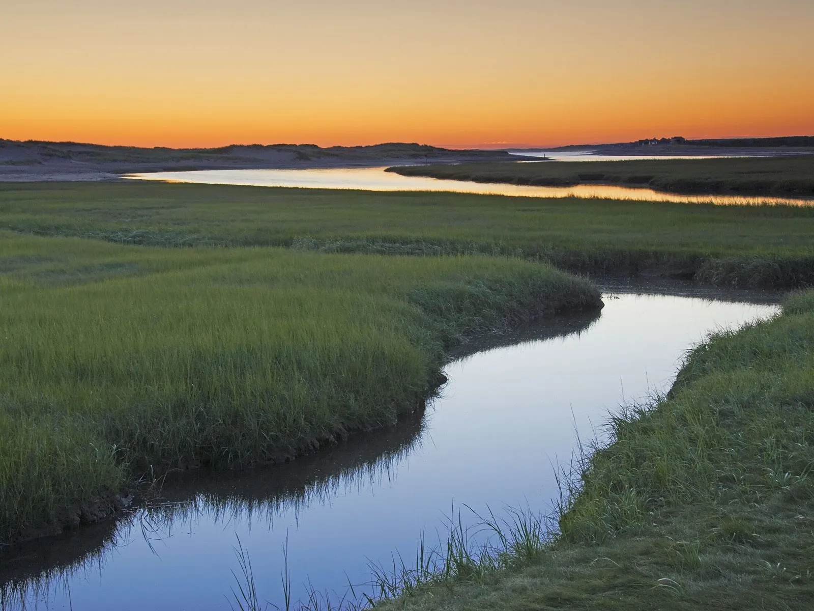 A calm river flows through a green marshland under a vibrant orange sunset sky.