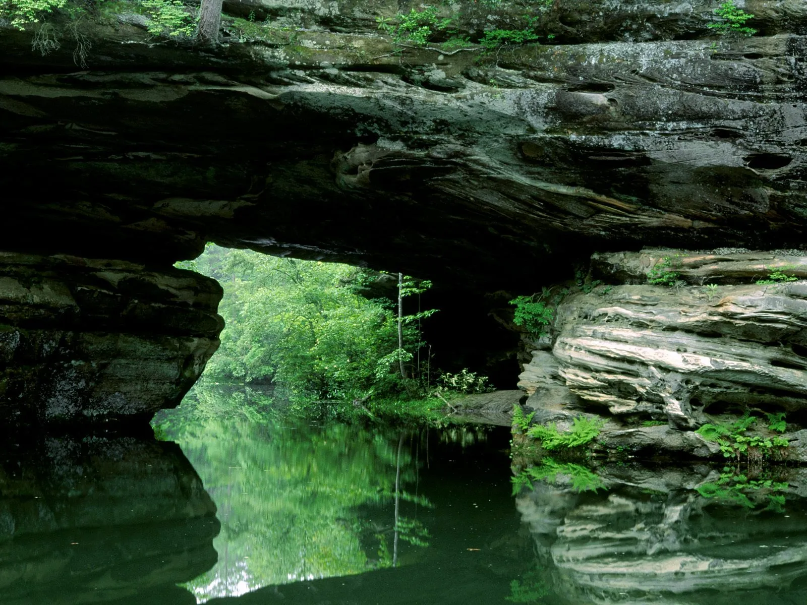 A calm river flows through a natural rock archway, with lush green trees reflected in the water.