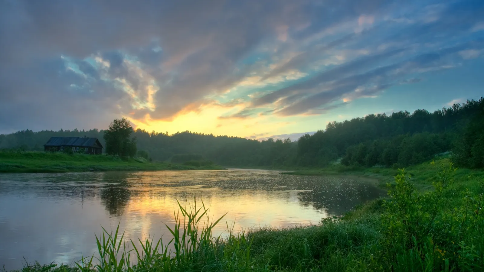 A wide river flows past a grassy bank with a small cabin and trees, under a colorful sunrise sky.