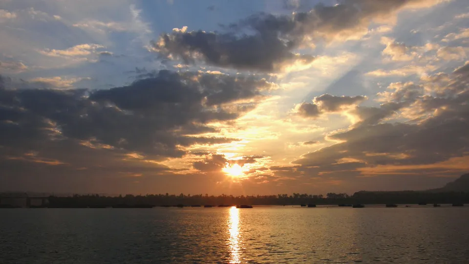 The sun sets over a wide river, casting golden rays through dramatic clouds and reflecting on the water.