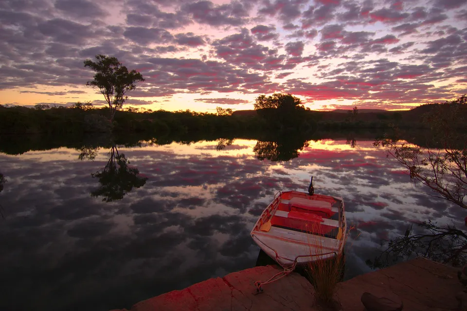 A small boat floats on a calm river reflecting a vibrant sunset sky with scattered clouds.