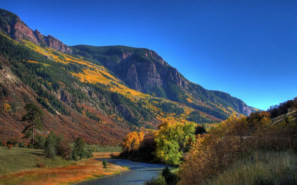 A river flows through a valley with mountainsides covered in yellow and orange autumn foliage under a blue sky.
