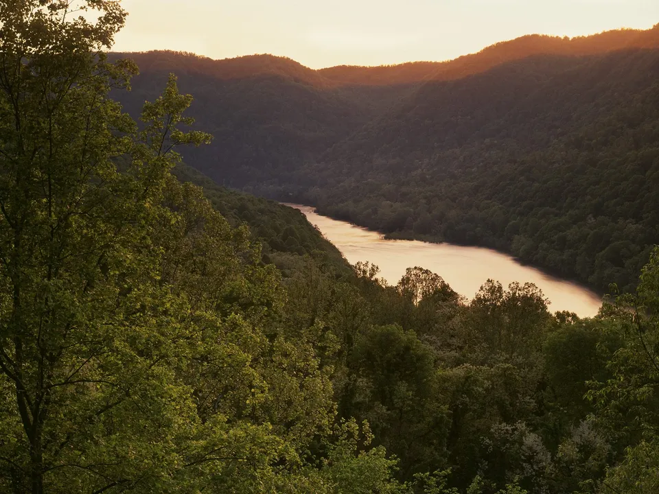 A wide river flows through a valley surrounded by lush green mountains, illuminated by the warm glow of sunset.