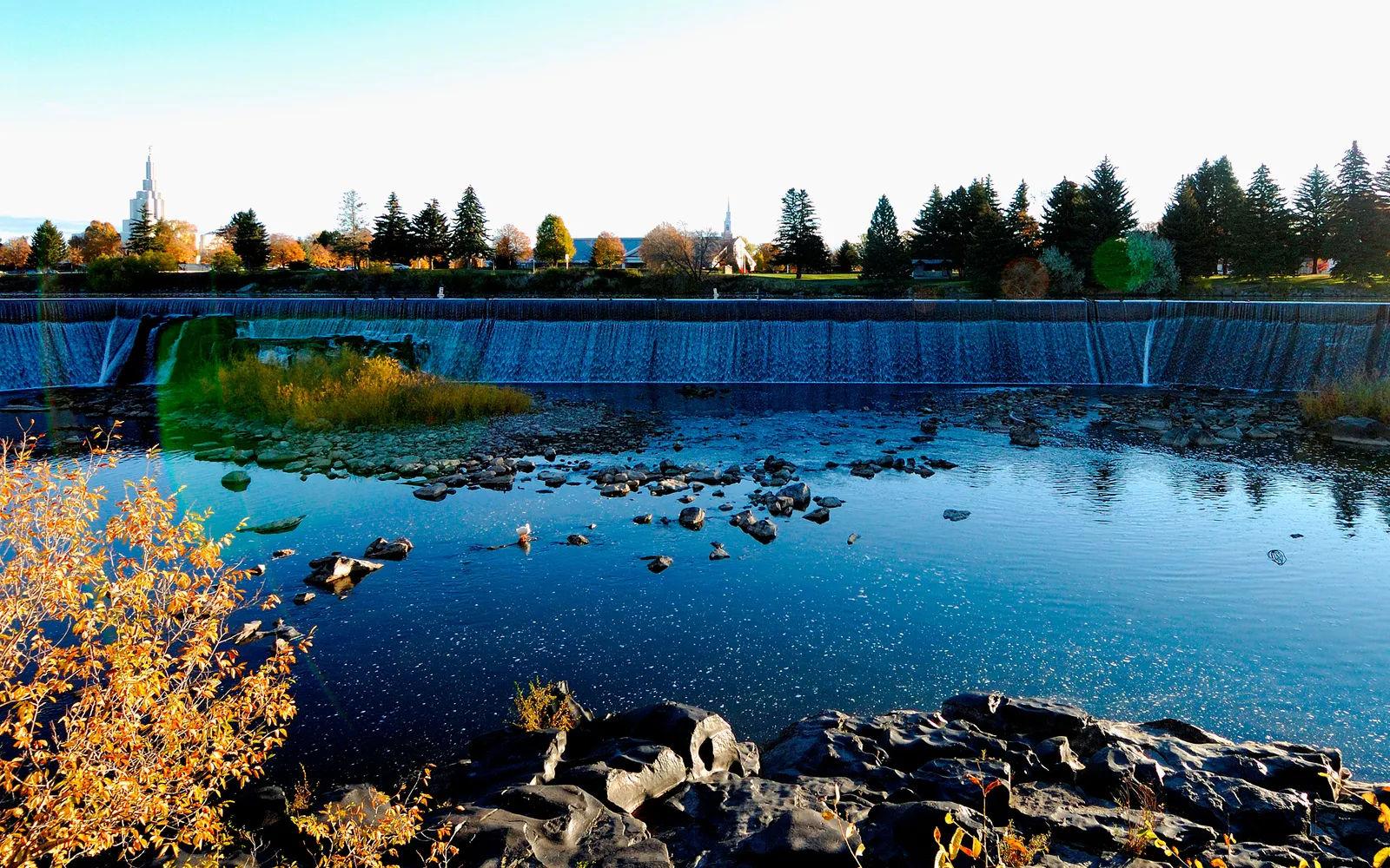 A wide river waterfall flows over a dam with autumn foliage in the foreground and buildings in the background.