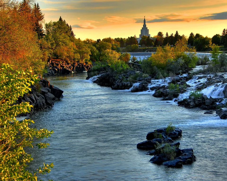 A river flows past rocky banks and small waterfalls towards a distant temple at sunset.