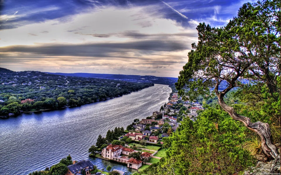 A wide river flows through a landscape dotted with houses and surrounded by trees, under a dramatic sky.