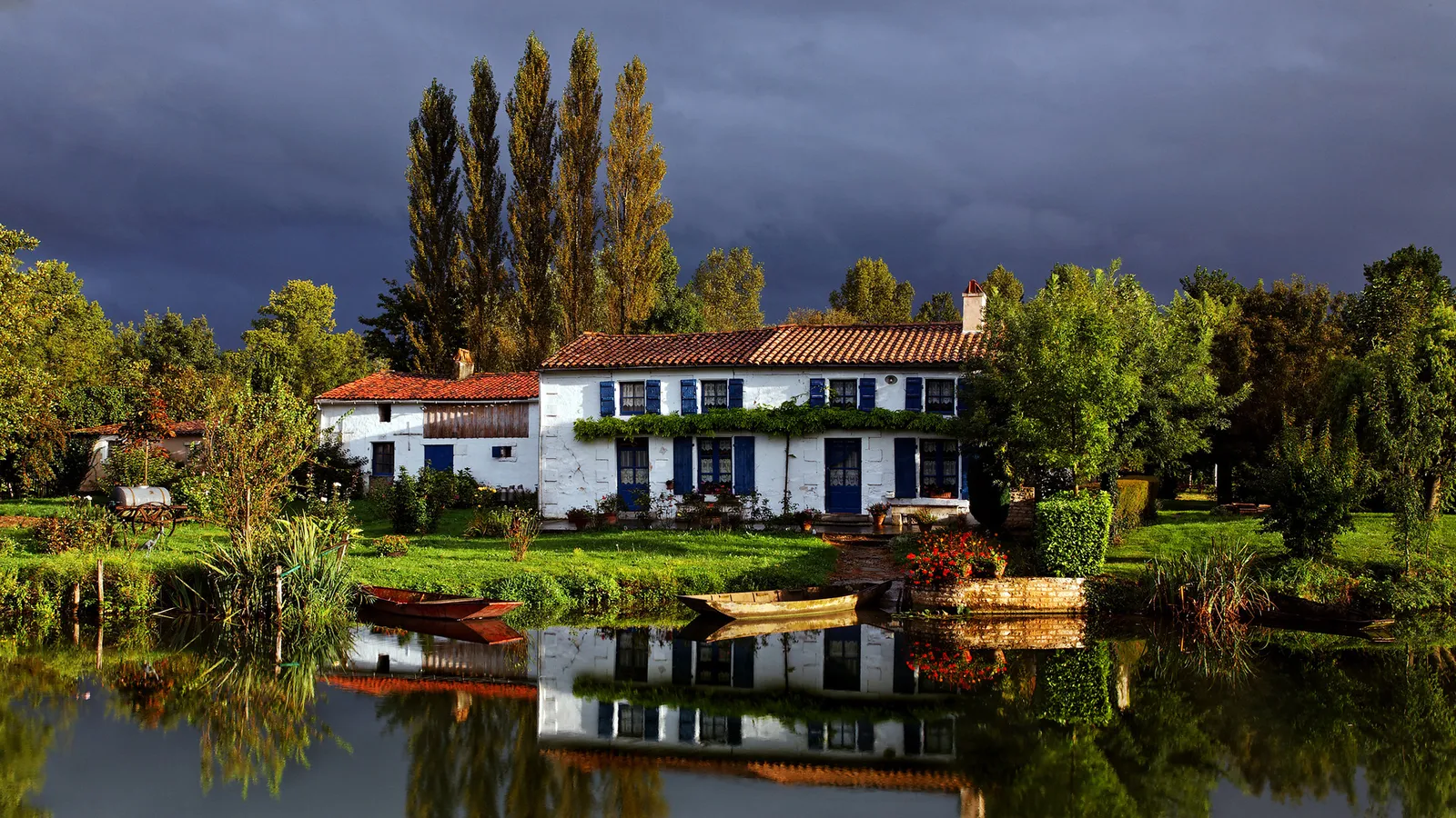 A white house with blue shutters sits beside a calm river, reflecting the trees and stormy sky.