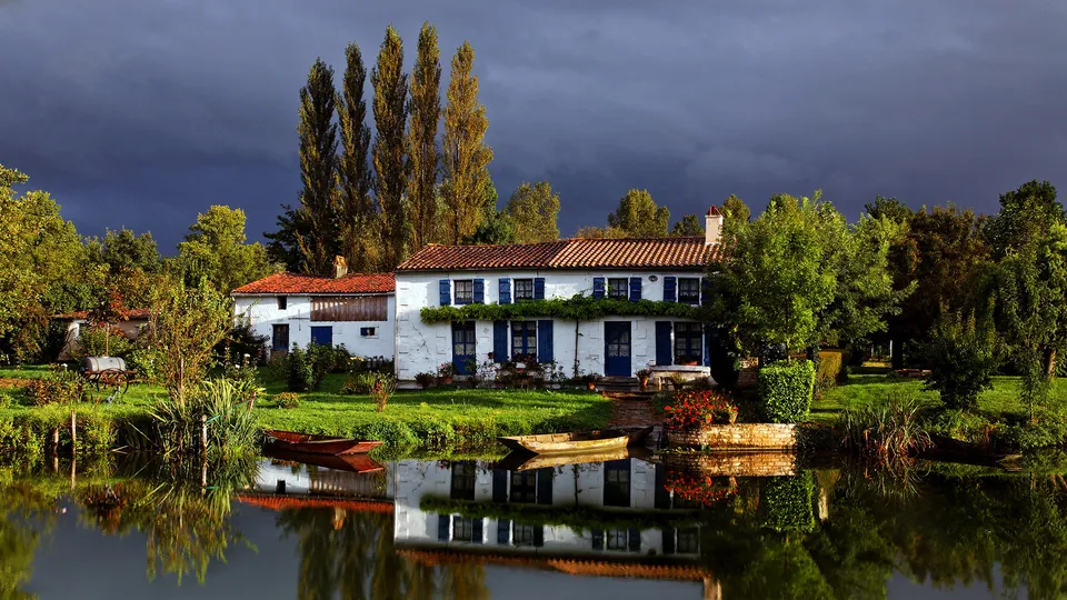 A white house with blue shutters sits beside a calm river, reflecting the trees and stormy sky.