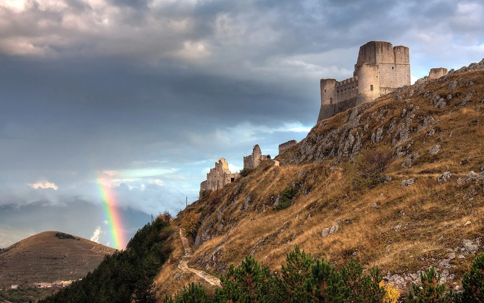 Ruins of a medieval castle perched on a rocky hillside with a rainbow in the stormy sky.