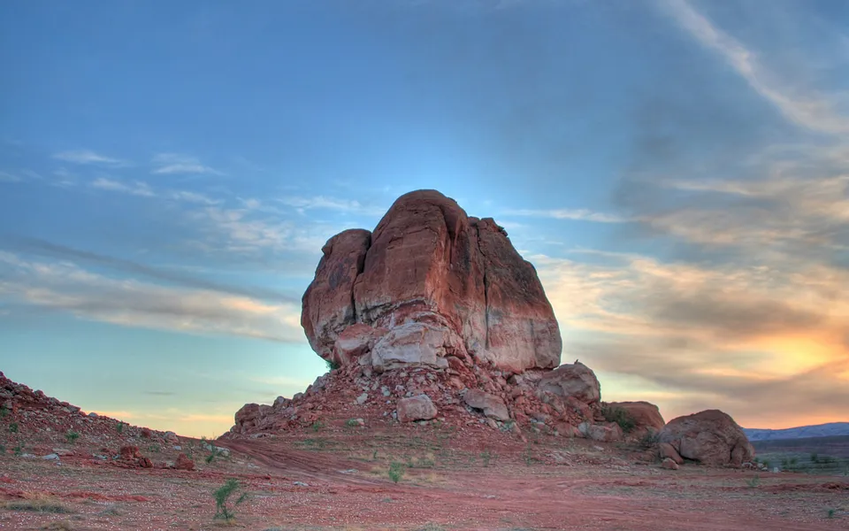 A large, reddish-brown rock formation stands in a desert landscape under a blue sky with wispy clouds.