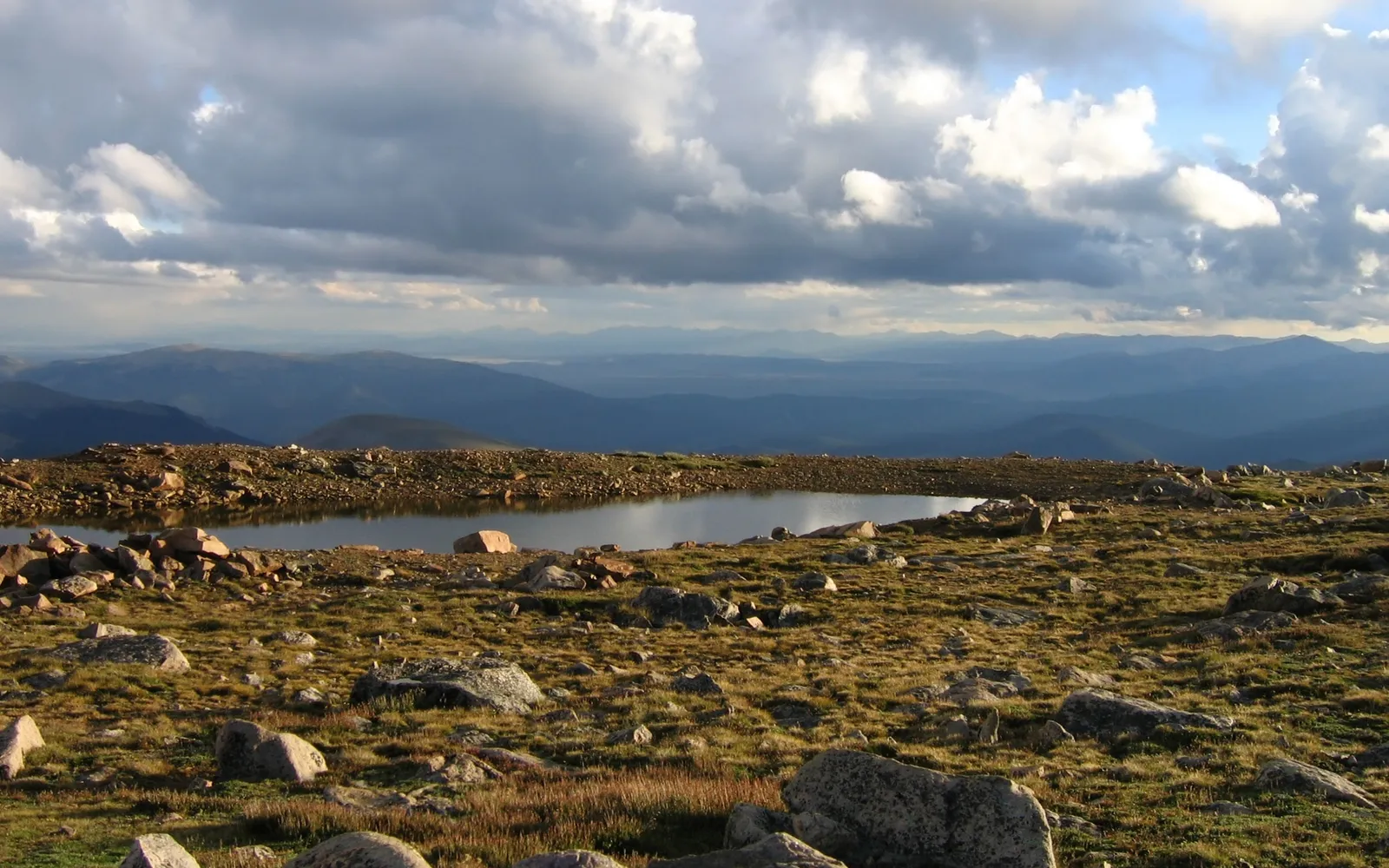 A calm alpine lake reflects a dramatic, cloudy sky, surrounded by rocky terrain and distant hazy mountains.