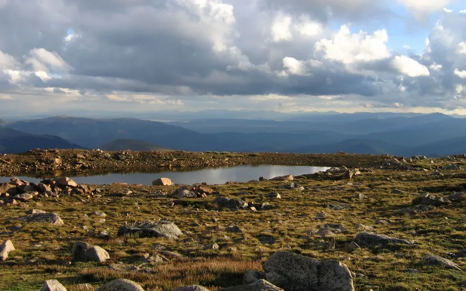 A calm alpine lake reflects a dramatic, cloudy sky, surrounded by rocky terrain and distant hazy mountains.