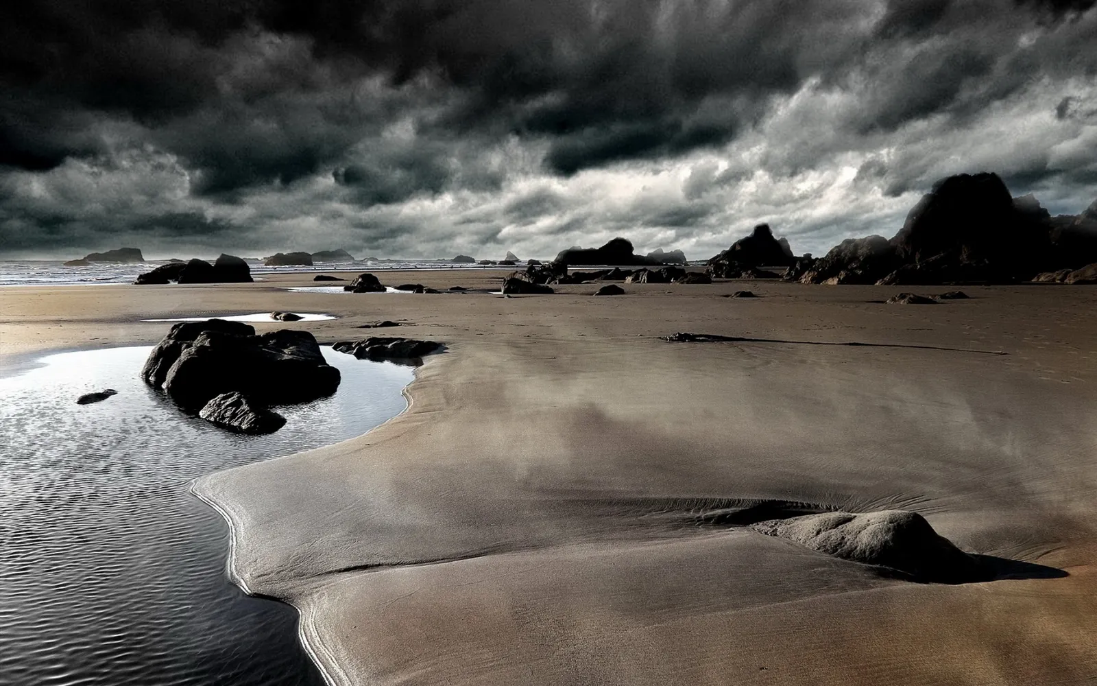 A wide, sandy beach with dark, stormy clouds overhead and scattered rocks.