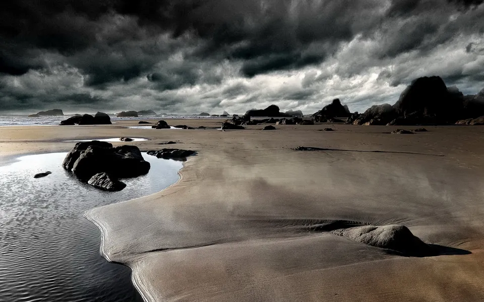 A wide, sandy beach with dark, stormy clouds overhead and scattered rocks.