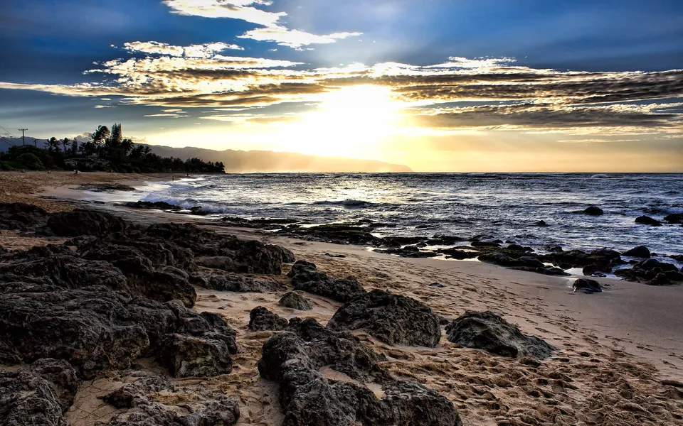 A dramatic sunset over a rocky beach with waves crashing on the shore and mountains in the distance.