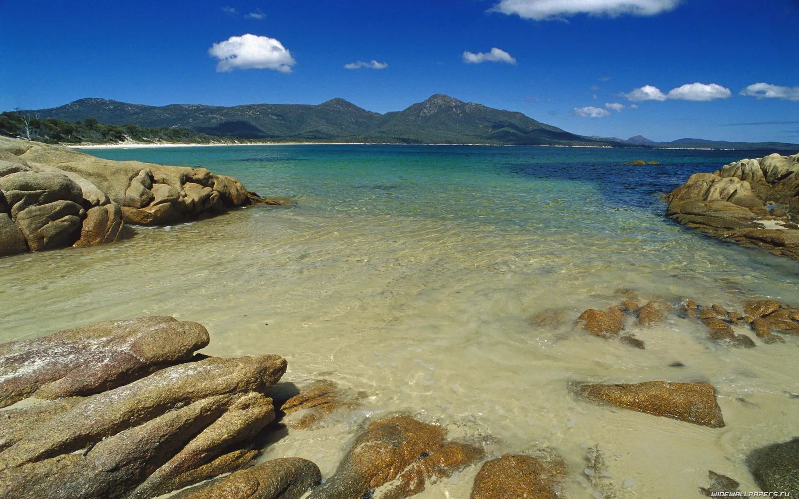 A clear turquoise ocean laps against a sandy beach with large, weathered rocks in the foreground and mountains in the distance.