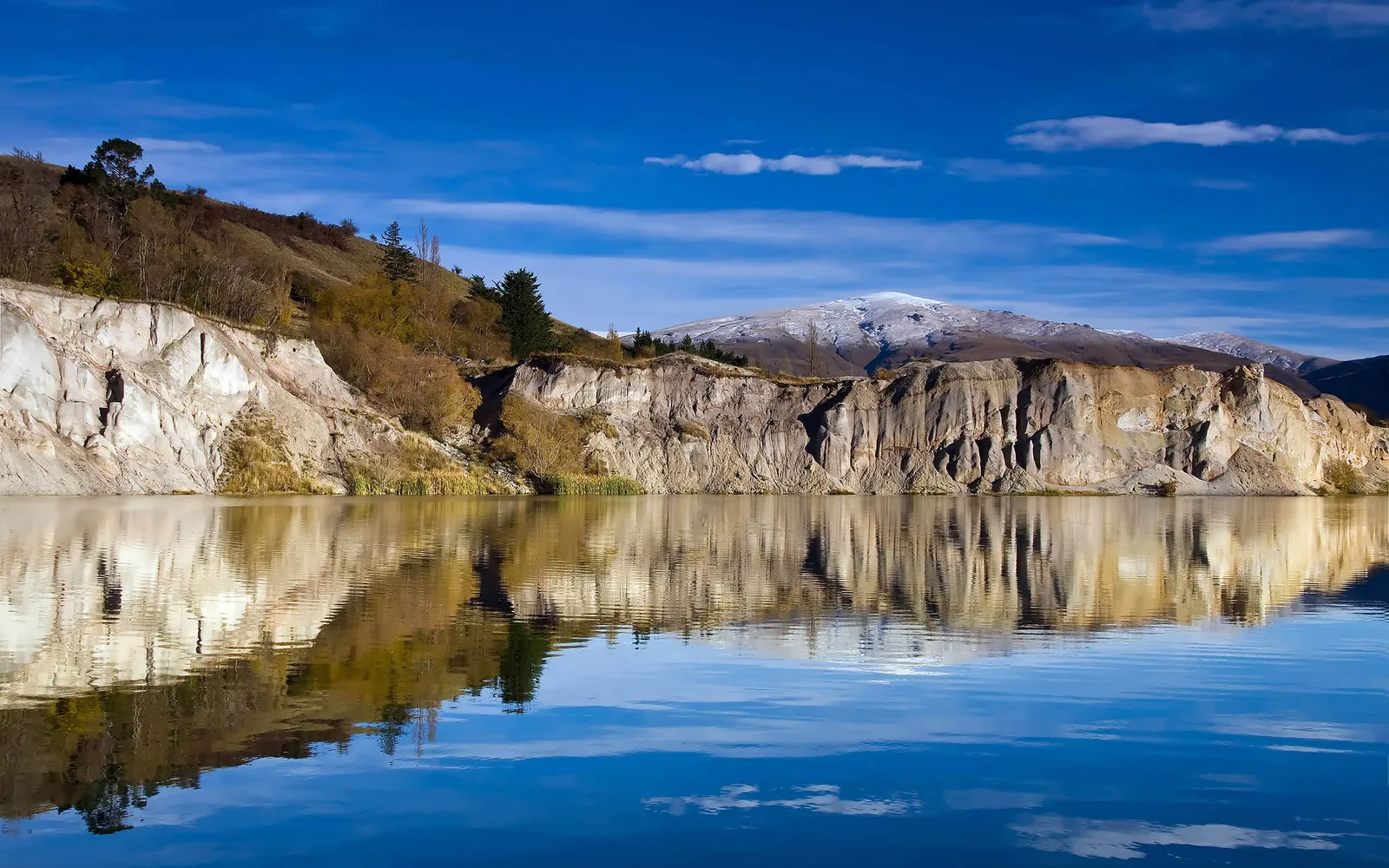 A calm lake reflects the textured cliffs and snow-capped mountains under a bright blue sky.