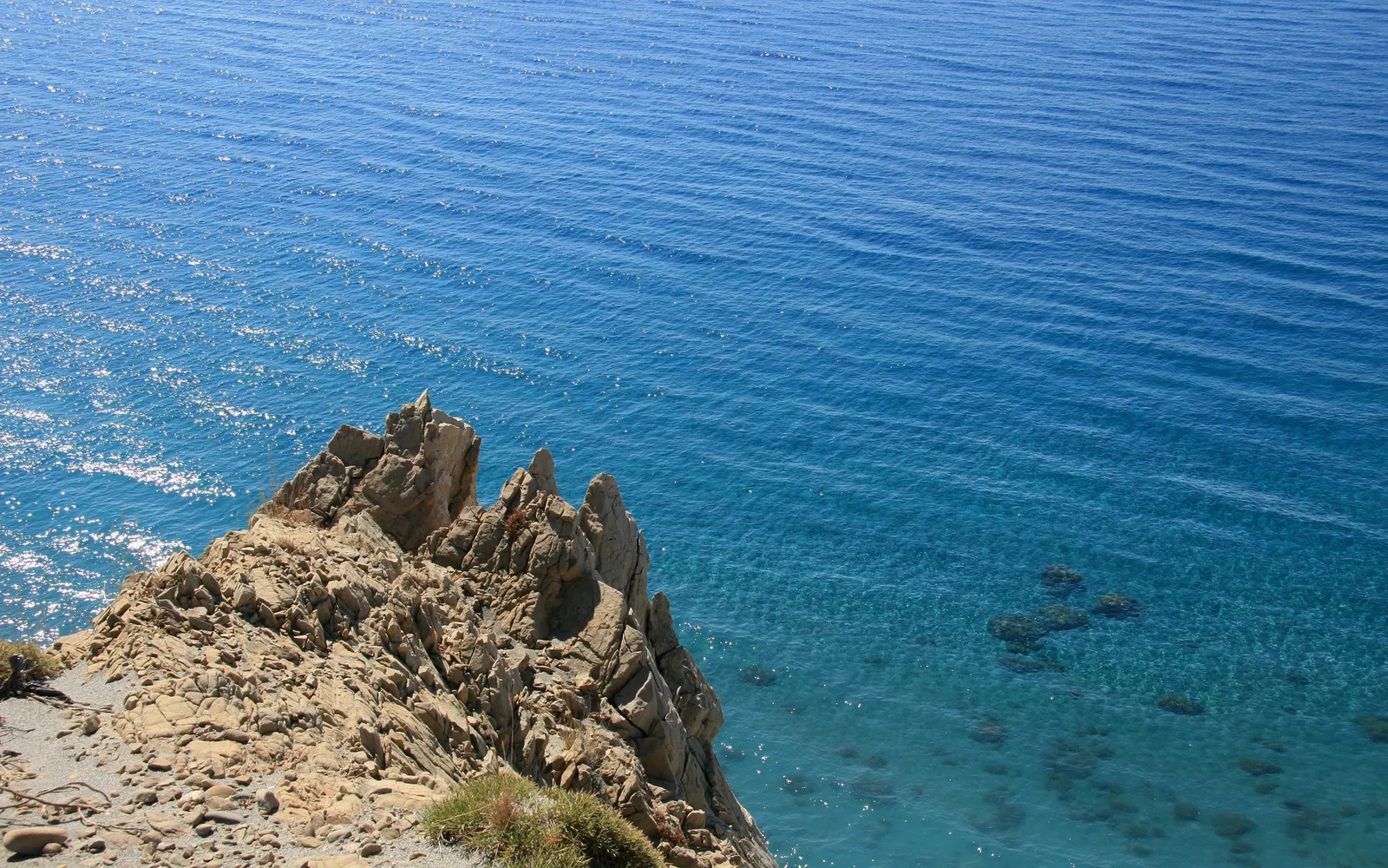 A rocky cliff face juts out into the bright blue ocean, with clear water revealing submerged rocks.