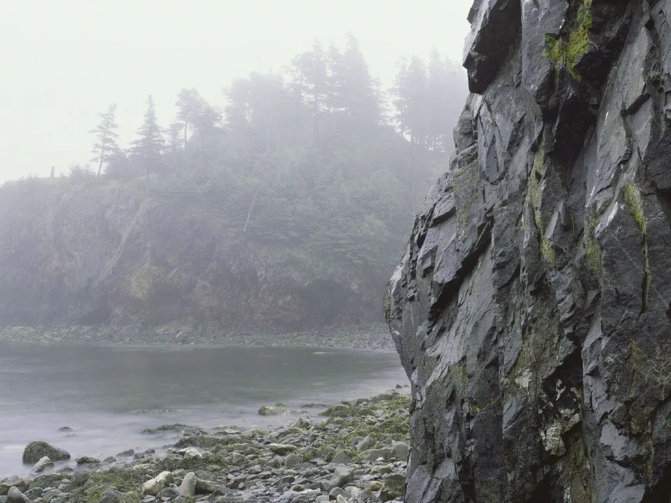 A rugged, dark rock face juts out into a misty, calm body of water with a forested hill in the background.