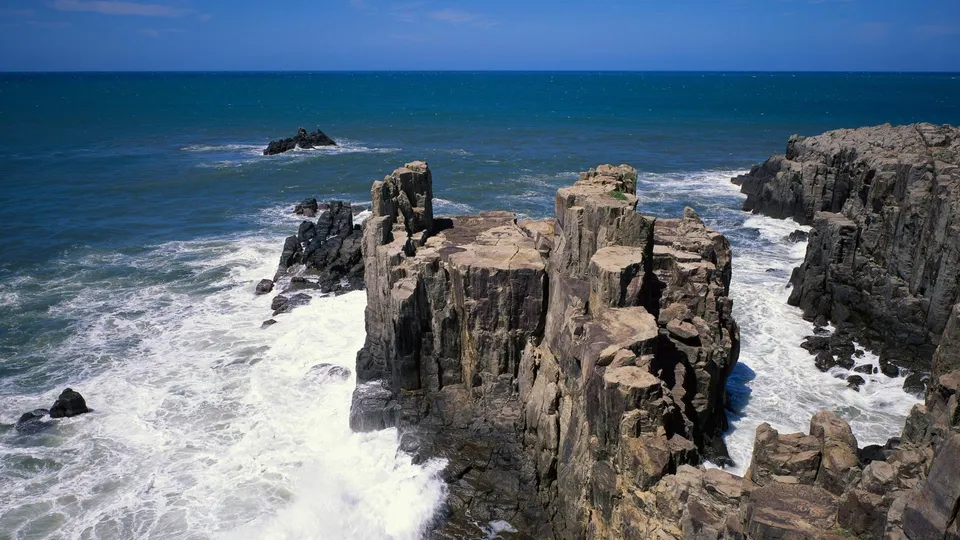 Jagged, dark rock cliffs rise from the turbulent blue ocean with white waves crashing against them.