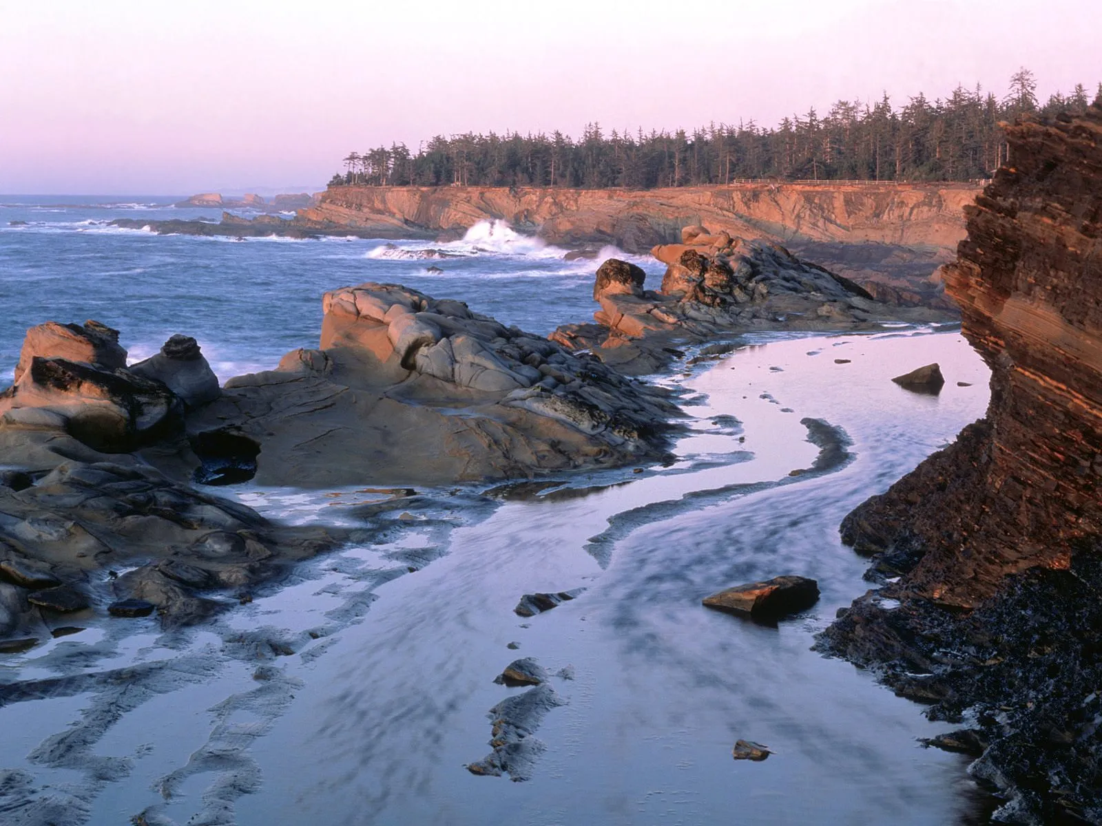 Waves crash against rugged rocks on a sandy beach during a soft sunset.