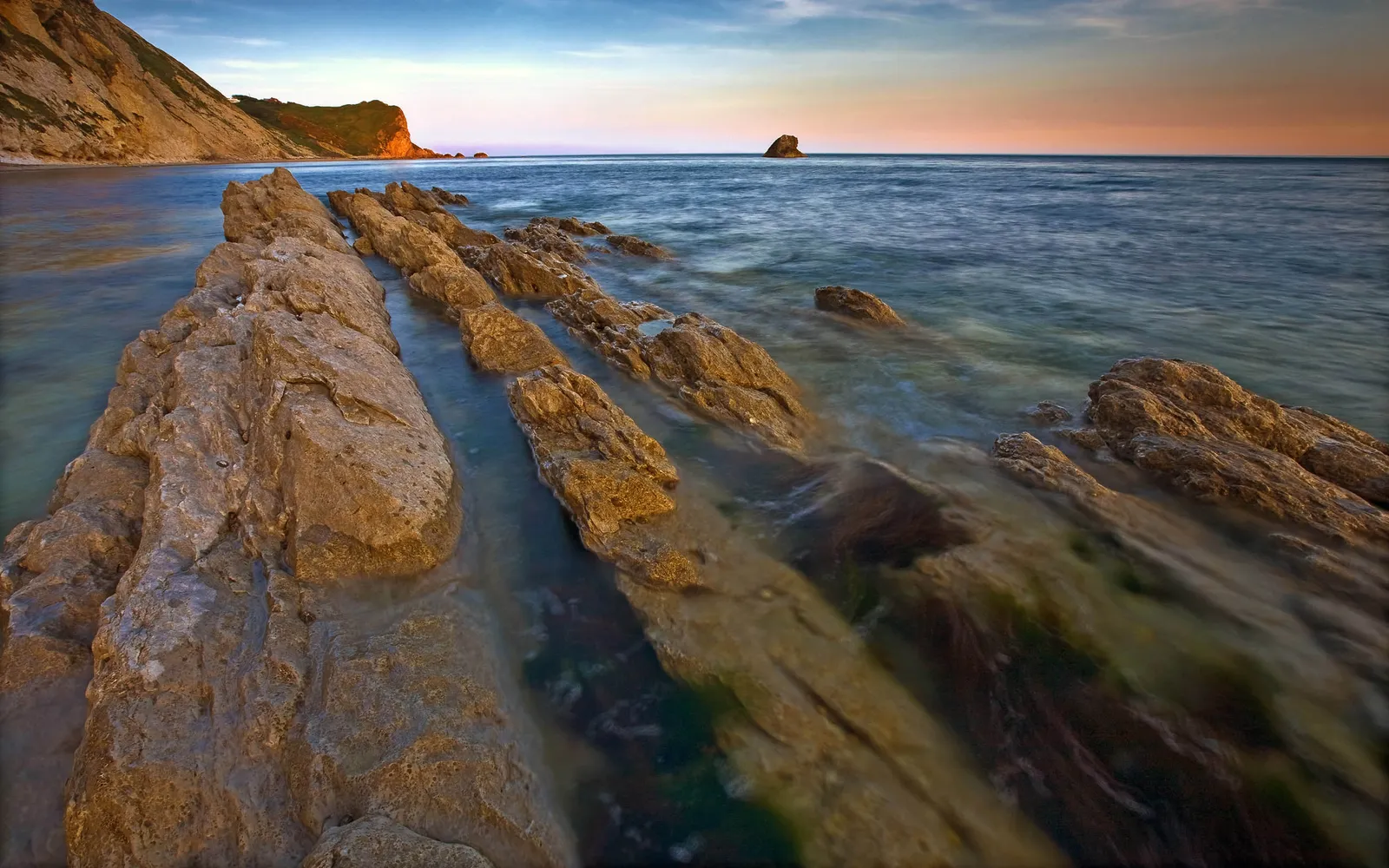 Long exposure of waves washing over layered rocks on a coastline at sunset.
