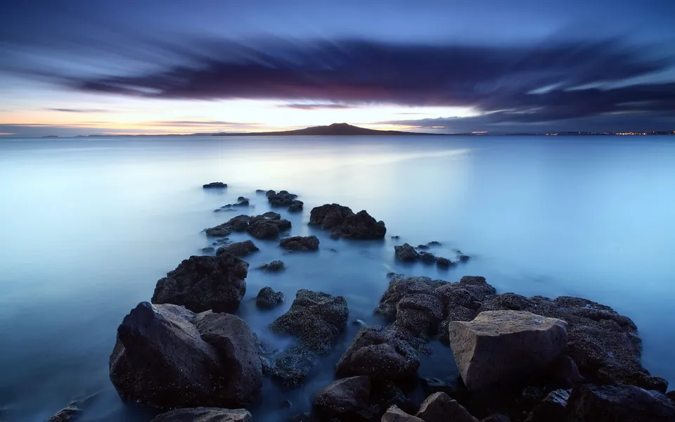Rocks emerge from the calm, misty water under a deep blue and purple sky at dusk.