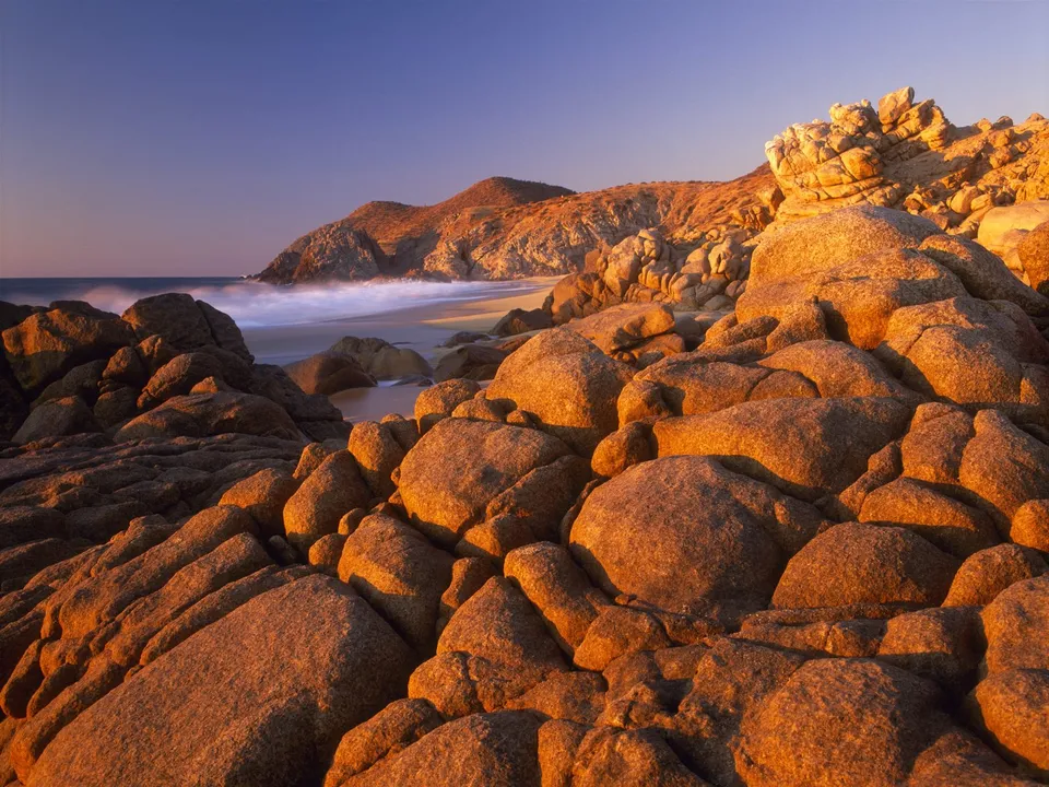 A rocky coastline bathed in the warm light of the setting sun, with waves crashing on the shore.