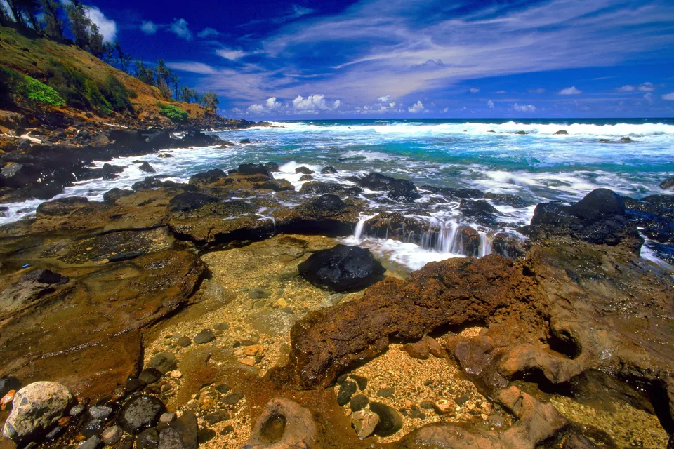 Waves crash against dark volcanic rocks and tide pools on a sunny coastline with a blue sky.