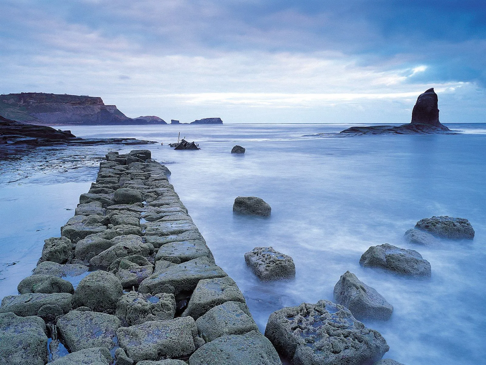 A stone path leads into the calm ocean with large rocks scattered in the water and cliffs in the distance.