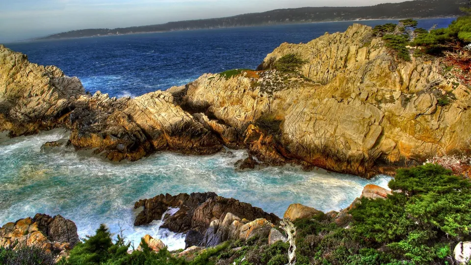 A rugged, rocky coastline with turquoise ocean waves crashing against the shore, under a hazy blue sky.