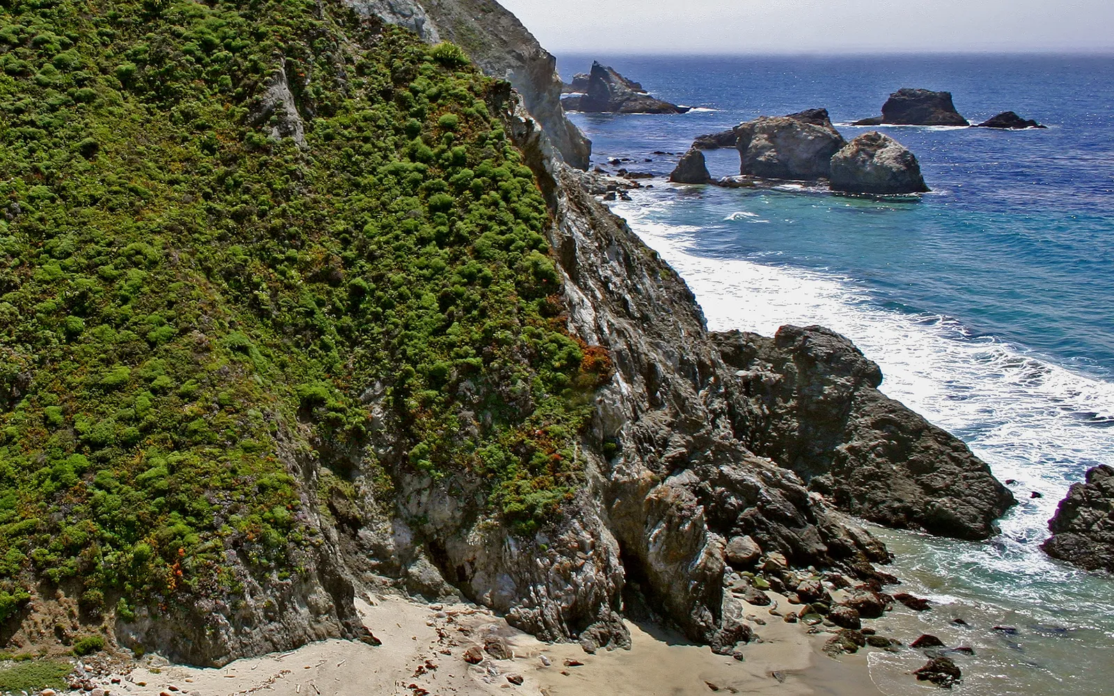 A steep, green-covered cliff drops to a sandy beach and the blue ocean with rock formations.