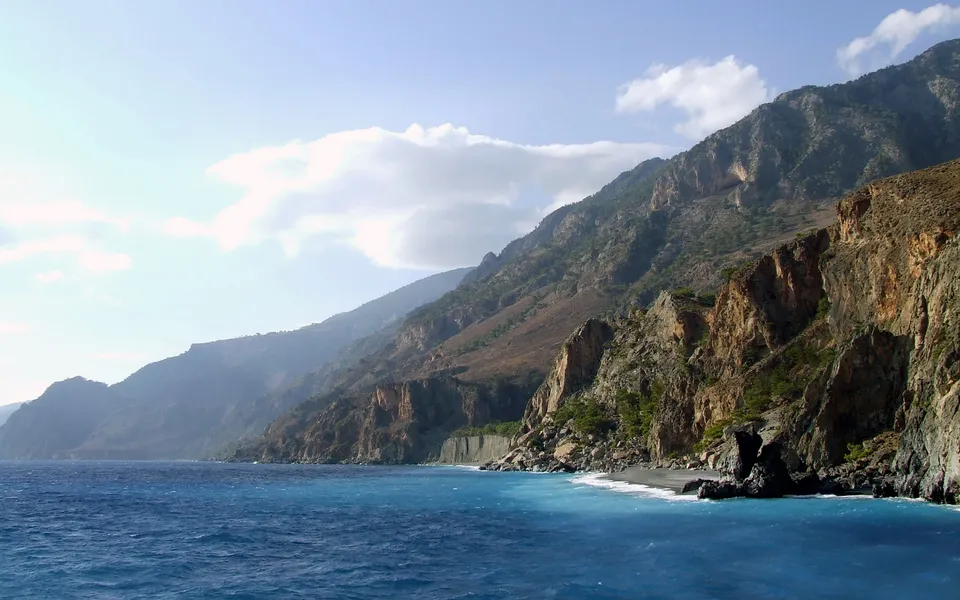 A rugged coastline with steep, rocky cliffs meeting the bright blue ocean under a partly cloudy sky.