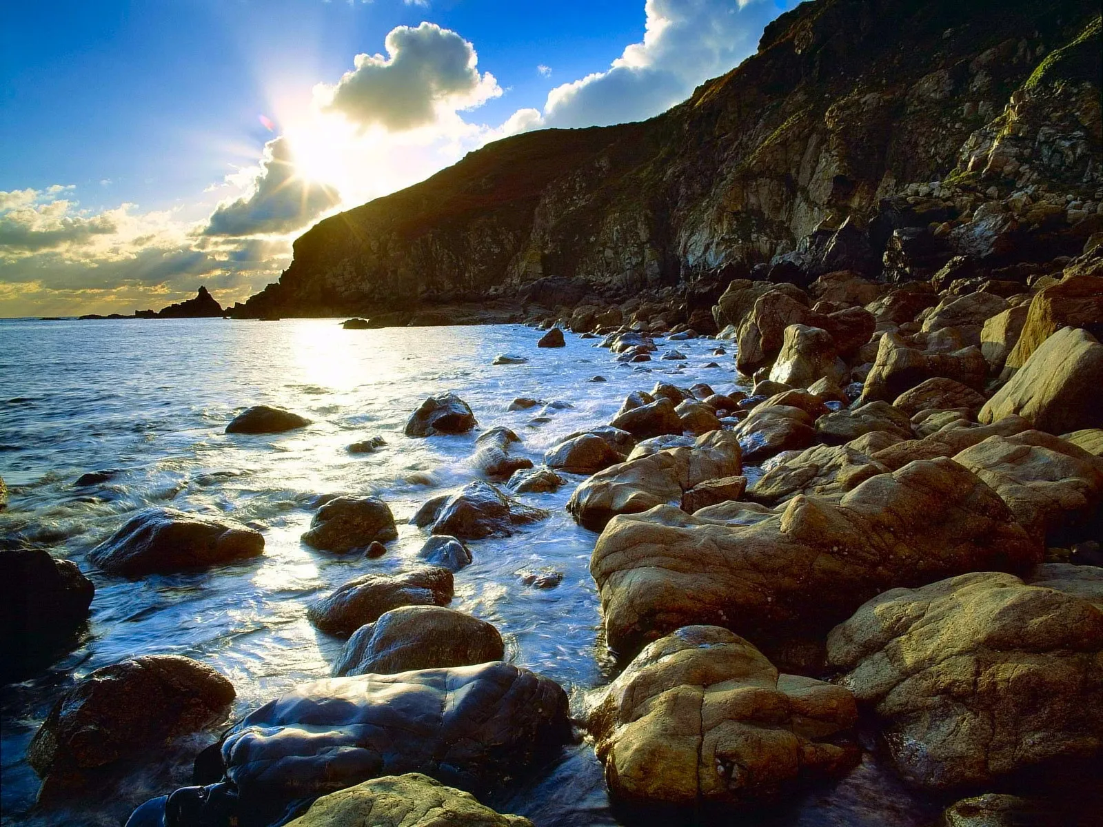 The sun shines brightly over a rocky coastline with waves gently lapping at the shore.
