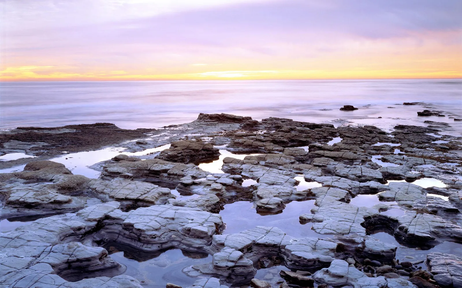 A pastel sunrise over the ocean, with tide pools reflecting the sky on a rocky shore.