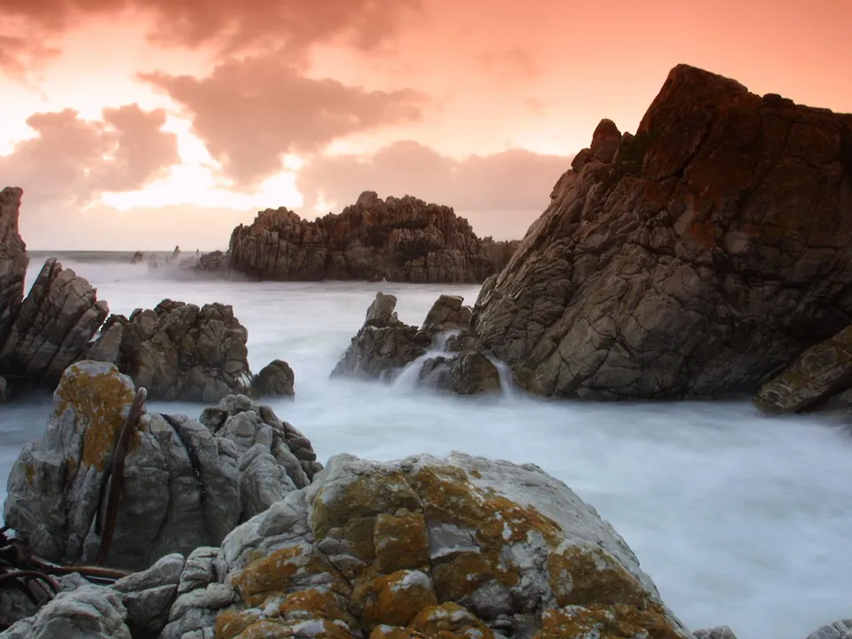 A dramatic sunset with orange clouds over a rocky coastline with waves crashing.