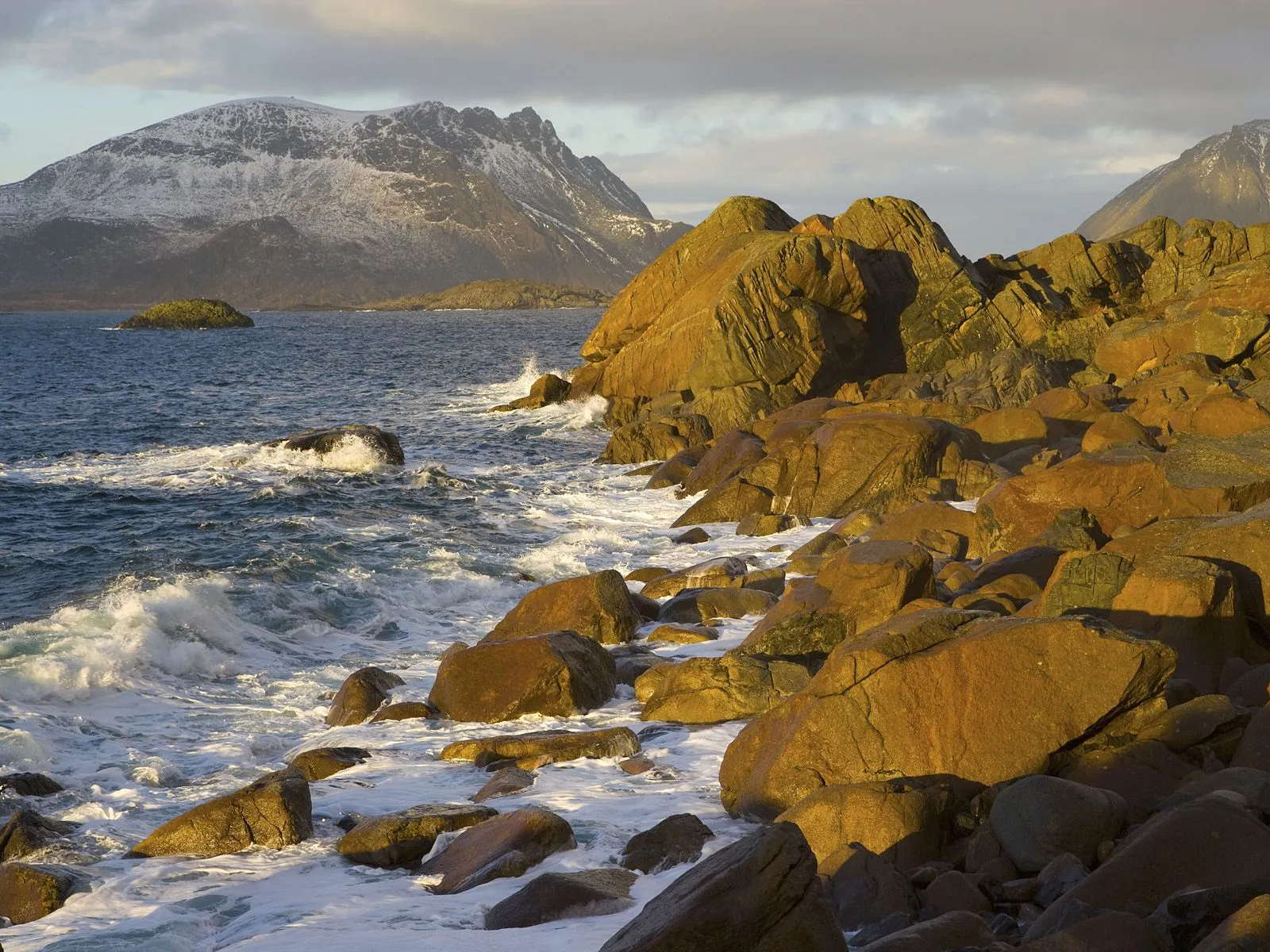 Waves crash onto a rocky shoreline with mountains in the background during sunset.