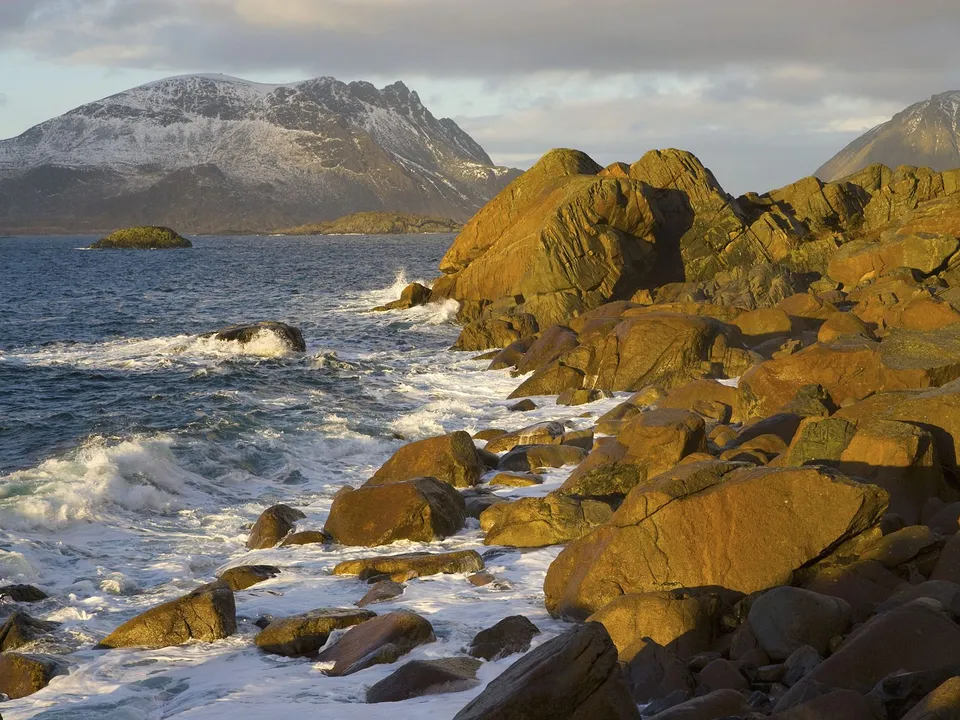 Waves crash onto a rocky shoreline with mountains in the background during sunset.