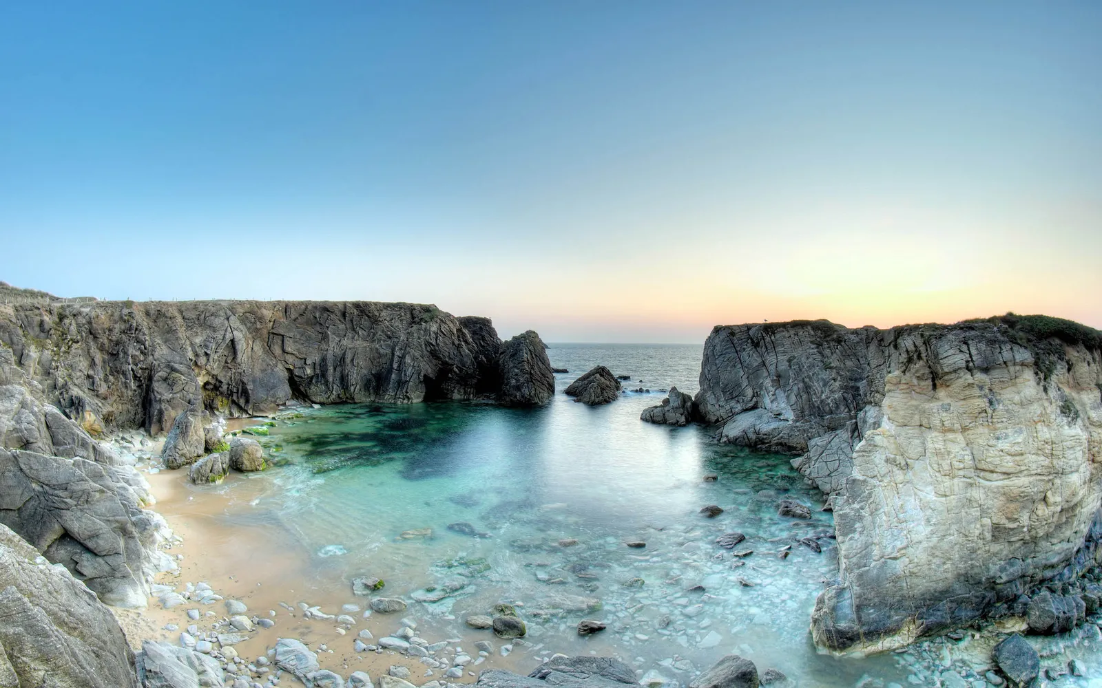 A panoramic view of a secluded rocky cove with clear turquoise water and dramatic cliffs under a pale sky.