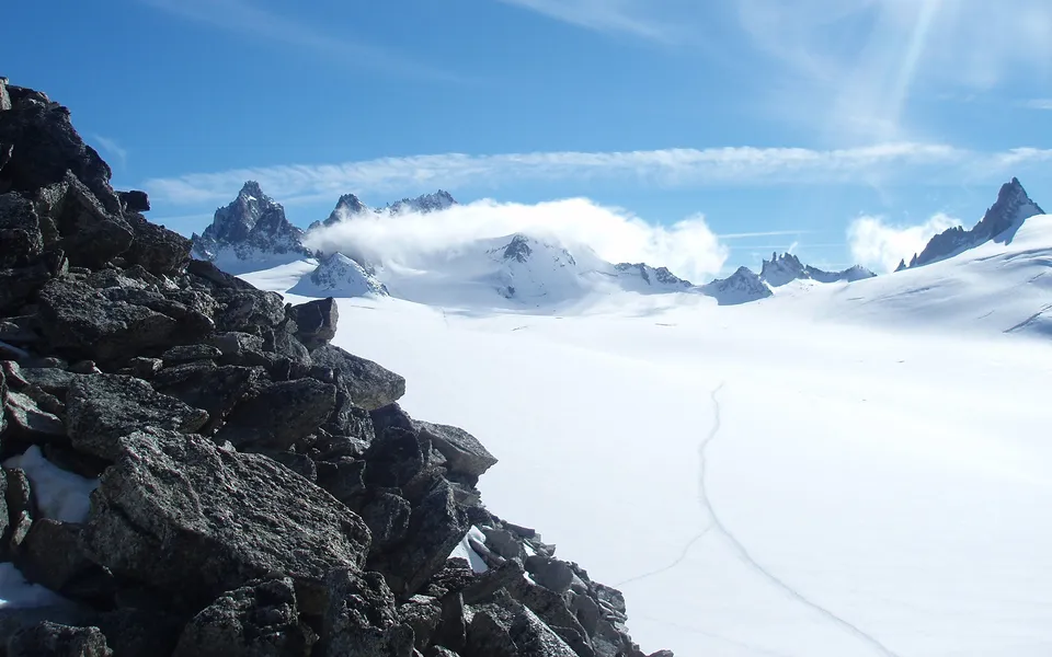 A dramatic mountain landscape with a rocky foreground, vast snowfields, and snow-covered peaks under a blue sky with clouds.