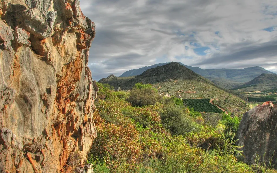 A close-up of a textured, orange-hued rock face in the foreground, with rolling green hills and mountains under a cloudy sky in the background.