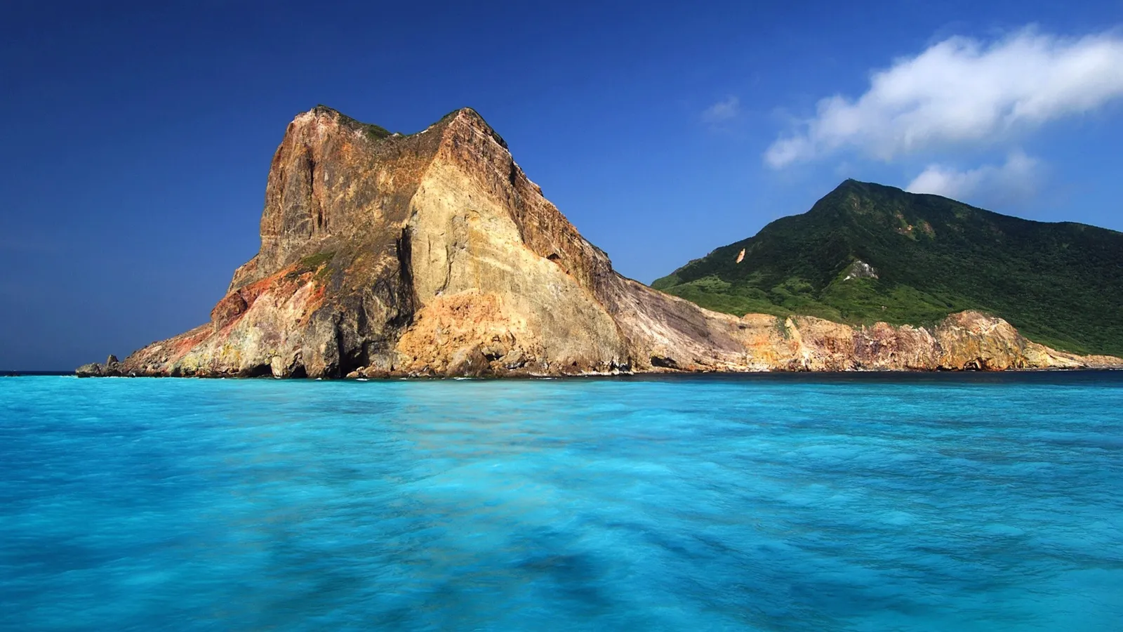 A rugged, multi-colored rocky island juts out of vibrant turquoise ocean water under a clear blue sky.