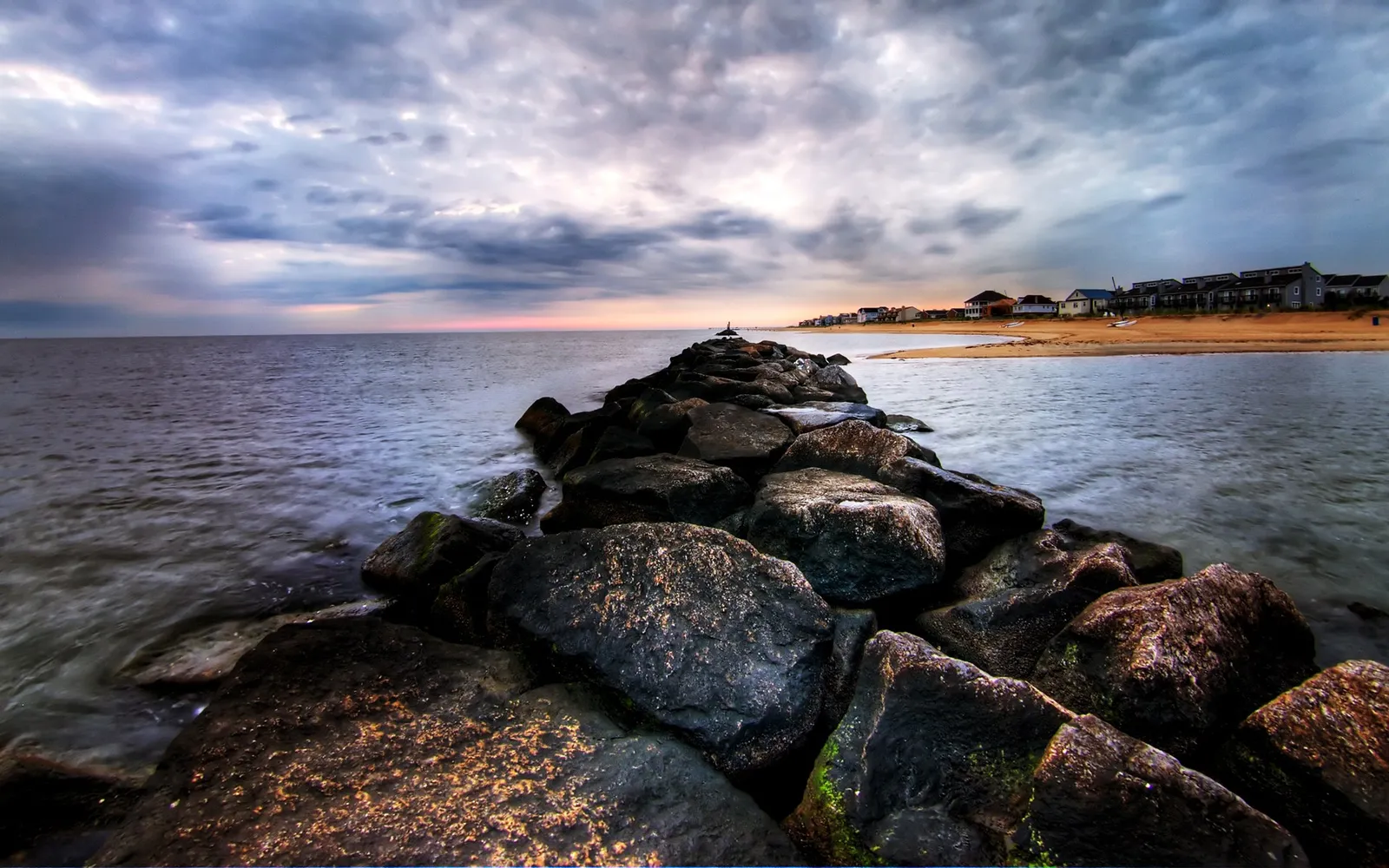 A long jetty made of large rocks extends into the ocean under a cloudy sky at sunset.