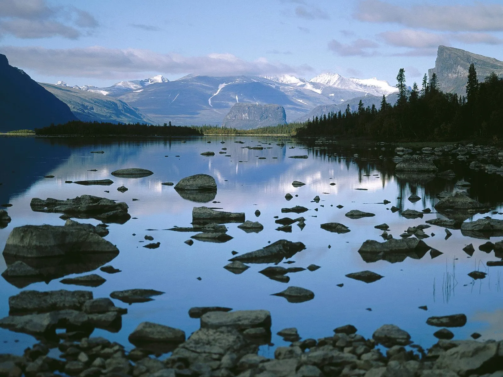 A calm lake with scattered rocks in the foreground, reflecting snow-capped mountains and a dark forest under a cloudy blue sky.