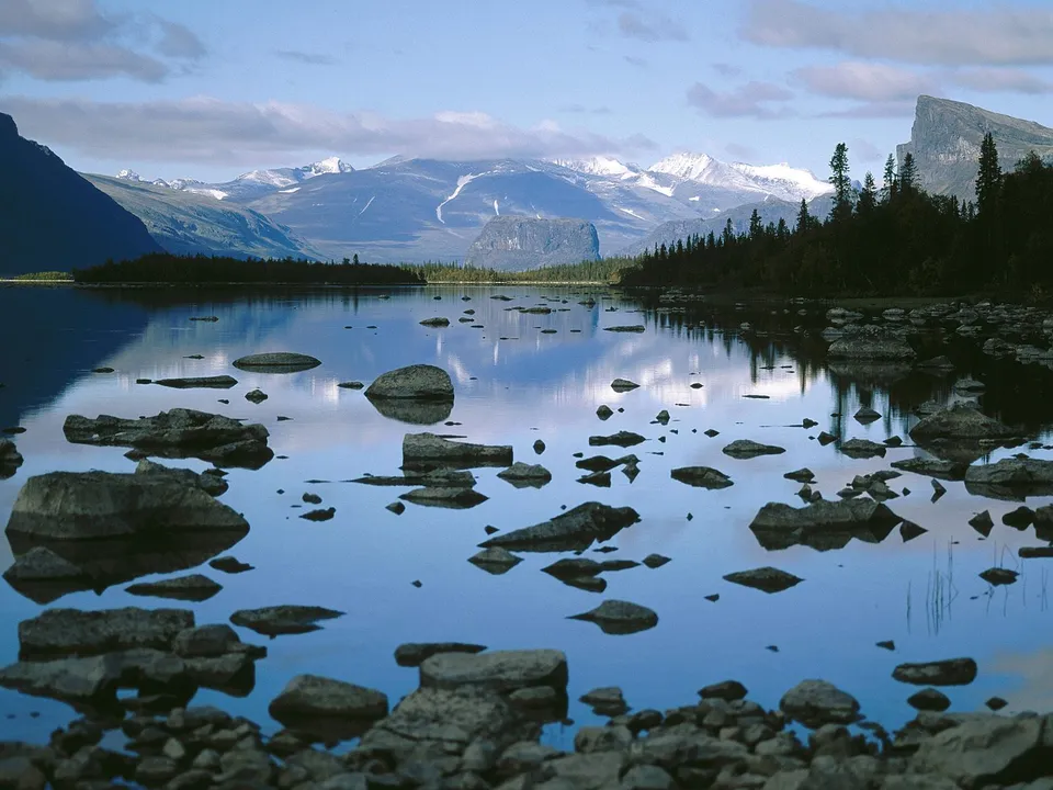 A calm lake with scattered rocks in the foreground, reflecting snow-capped mountains and a dark forest under a cloudy blue sky.