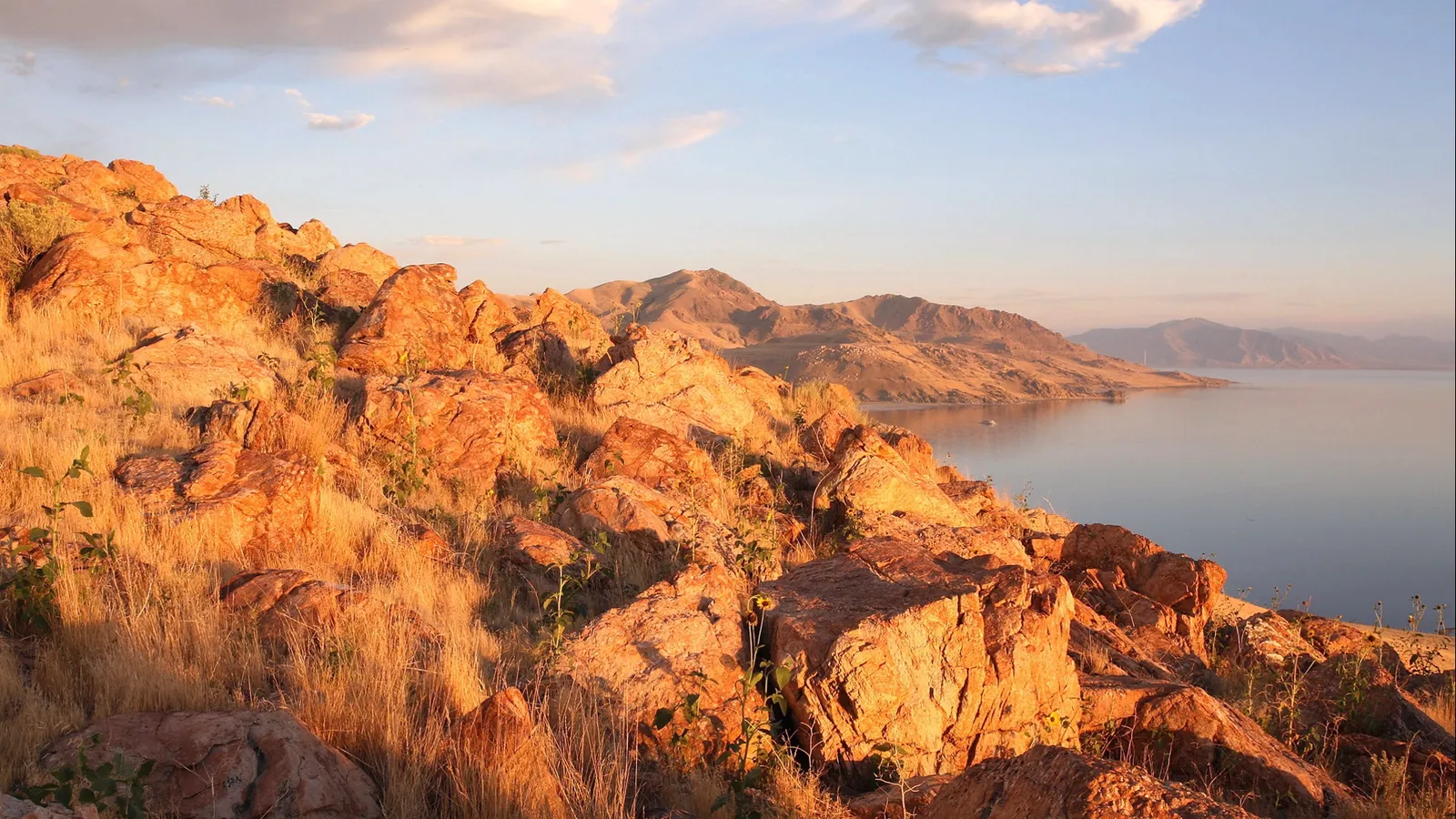 A rocky shoreline bathed in golden hour light, overlooking a calm lake and distant mountains.