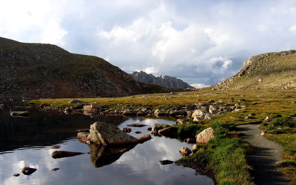 A mountain lake reflects rocky terrain and cloudy skies with a path in the foreground.